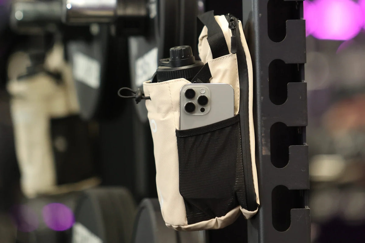 Beige RYVO The Rig magnetic gym bag stuck to a steel squat rack upright, with a phone in the front pocket and a drink bottle up top, kept off the gym floor.