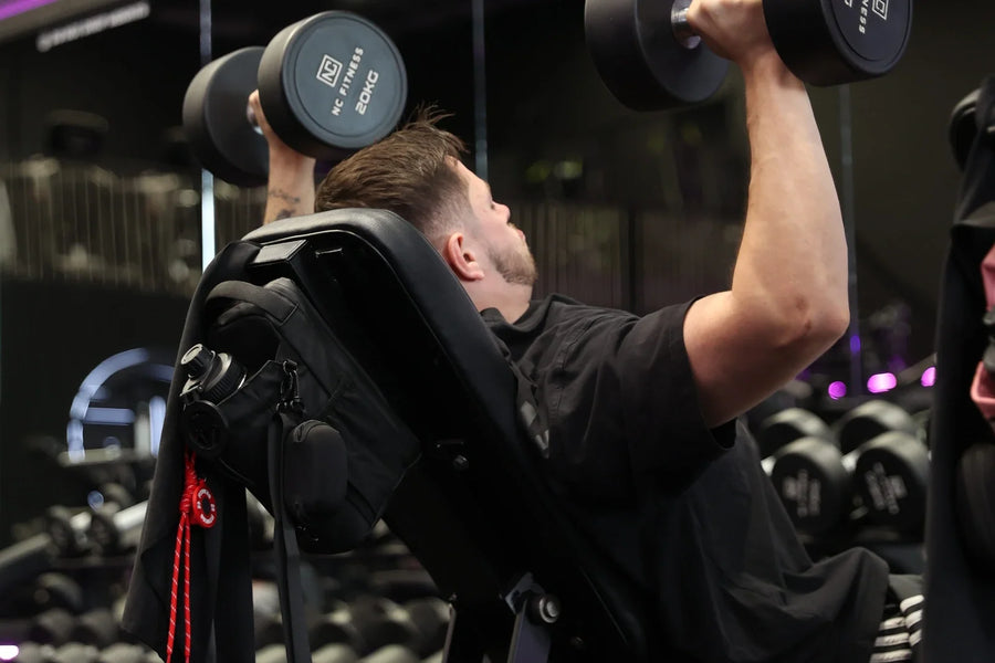Man mid-set on an incline dumbbell press, focused on the lift, with a black RYVO The Rig Pro gym bag holding his bottle and earbud case on the back of the bench, in a gym lit with purple lights.