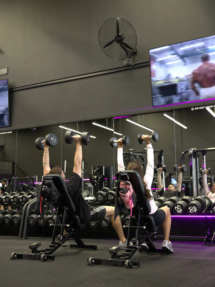 Two people doing shoulder presses at the gym with RYVO gym bags nearby